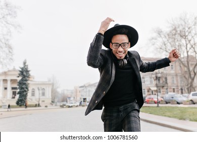 Spectacular african man in glasses running in autumn park. Outdoor portrait of stylish black guy in hat fooling around in city square.