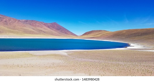 Spectacular Contrast Between The Miscanti Lagoon With Its Prussian Blue Waters And The Pink Slopes Of Miñiques Volcano, Near San Pedro De Atacama, Chile.