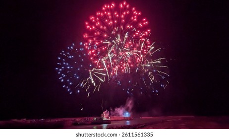 Spectacular fireworks explode over the water, painting the night sky with vibrant reds, blues, and whites, creating a breathtaking display of light and color.