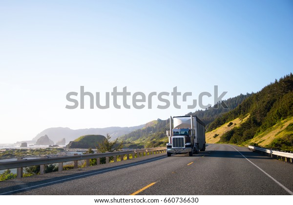 Spectacular landscaping with big rig semi truck and\
trailer which moving by empty highway along the Pacific coast with\
big cliffs in ocean waves and among the green hills on another\
side