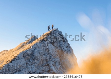 Spectacular view of two hikers walking along the mountain ridge route with the sun on the clear blue sky, aerial shot. Inspiration, nature, and mountaineering concepts.