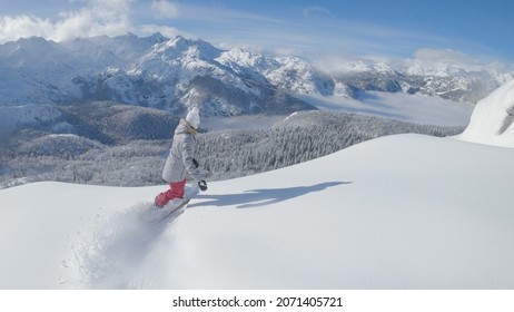 Spectacular view of the wintry landscape as young woman snowboards off-piste on a sunny day in the Julian Alps. Female traveler shreds the fresh powder snow while on a snowboarding trip in Slovenia.