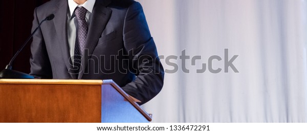 Speech of an abstract\
man in a suit on stage at the stand for performances. Tribune or\
pulpit for speaker official, president or professor. Close-up. Copy\
space.