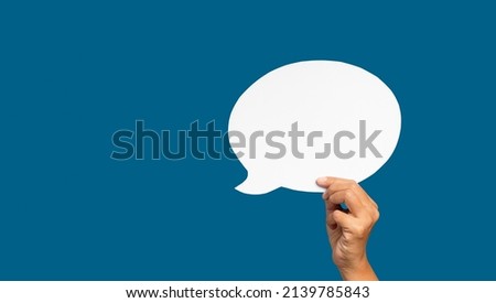 A speech bubble concept. Hand holding of an empty white speech bubble against a blue background. Space for text. Close-up photo.