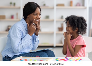 Speech Therapist Beautiful Young African American Woman Working With Little Black Girl With Curly Hair At Clinic, Sitting In Front Of Each Other, Smiling And Touching Their Faces, Side View