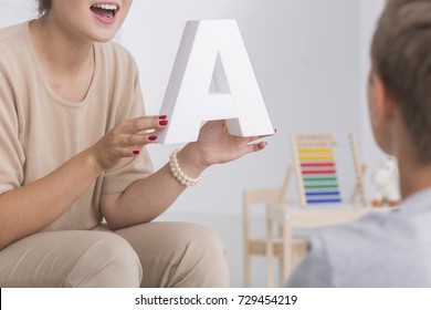 Speech Therapist In Beige Costume Holds Letter A And Speaks To Kid During Class Session