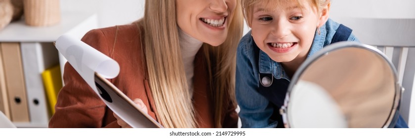 Speech Therapist Holding Clipboard Near Smiling Child And Mirror In Classroom, Banner