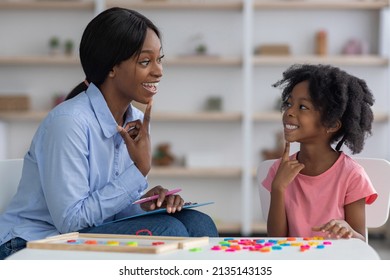 Speech Therapist Pretty Young African American Woman Working With Little Black Girl With Bushy Hair At Clinic, Sitting In Front Of Each Other, Smiling And Touching Their Chins, Side View