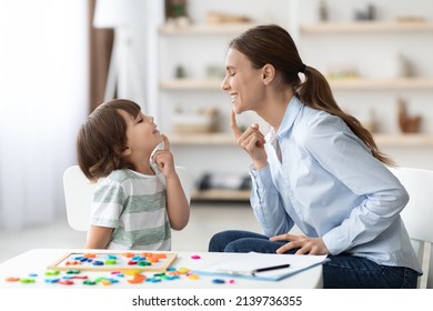 Speech training for kids. Professional woman specialist training with little boy at cabinet, teaching him right articulation exercises, side view