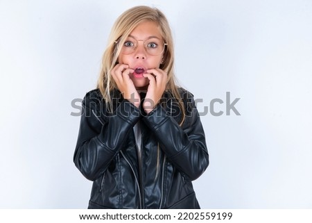 Speechless beautiful caucasian blonde little girl wearing biker jacket and glasses over white background keeps hands near opened mouth reacts to shocking news stares wondered at camera