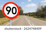 Speed limit sign on empty country road. Rural highway with 90 sign. Sunny day with blue sky. Asphalt road with white line. Road safety and traffic control. Transportation and legal regulation.