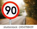 Speed limit sign with trees behind. Maximum ninety kilometers per hour. Safety on road background. White round sign red border line. Slow down foggy day. Autumn leaves road.