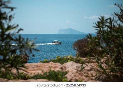 A speedboat glides across turquoise waters near a rocky coastline, framed by lush greenery and a distant island under a clear blue sky. - Powered by Shutterstock