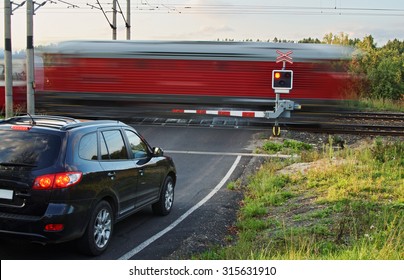 Speeding motion blur red train passing through a railway crossing with gates. Black car standing in front of the railway barriers on an asphalt road.