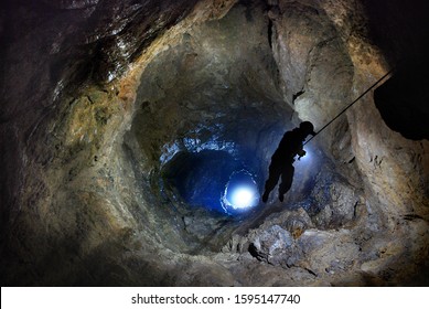 Speleologist descend by the rope in the deep vertical cave tunnel.  Cave man hanging over abyss. View from the top og the tunnel. Silhouette image. Karst cave. Caucasian mountains. Abkhazia. - Shutterstock ID 1595147740