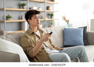 Spending Free Time. Portrait Of Asian Guy Watching TV Show Or Film, Holding Remote Control, Switching Cable Channels. Young Man Sitting On Couch At Home In Living Room, Selective Focus