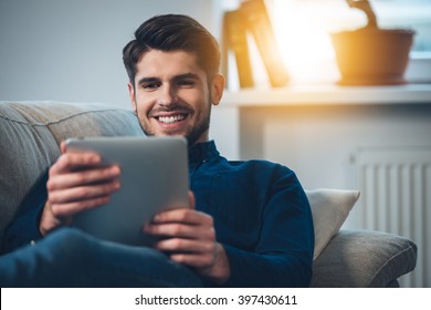 Spending Great Time At Home. Close-up Of Handsome Young Man Using His Digital Tablet With Smile While Lying Down On The Couch At Home