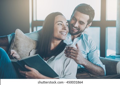 Spending nice time at home. Beautiful young loving couple bonding to each other and smiling while woman holding a book - Powered by Shutterstock