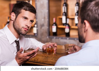 Spending Night In Bar. Two Confident Young Men In Shirt And Tie Talking To Each Other And Gesturing While Drinking Whisky At The Bar Counter 