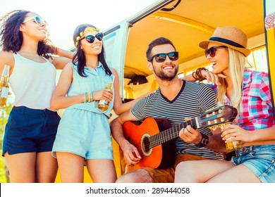 Spending Quality Time With Friends. Handsome Young Man Sitting At Minivan And Playing Guitar While Three Girls Standing Close To Him And Smiling 