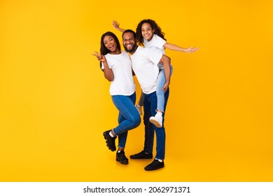 Spending Time With Family Is Fun. Full Body Length Of Excited African American Man, Woman And Girl Laughing And Posing Isolated Over Yellow Orange Studio Wall. Cheerful Dad Carrying Daughter On Back