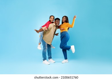 Spending Time With Family Is Fun. Full Body Length Of Excited African American Man, Woman And Girl Laughing And Posing Isolated Over Blue Studio Wall. Cheerful Father Carrying Daughter On Back, Banner