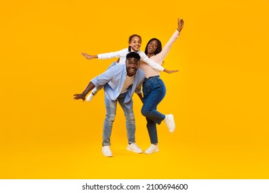 Spending Time With Family Is Fun. Full body length of excited African American man, woman and girl laughing and posing isolated on yellow studio wall. Cheerful father carrying daughter on back, banner - Shutterstock ID 2100694600