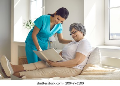 Spending time in a nursing home. Female caregiver in a nursing home discusses a book that an older woman is reading while lying in bed. Happy senior retired woman lying in bed and reading a book aloud
