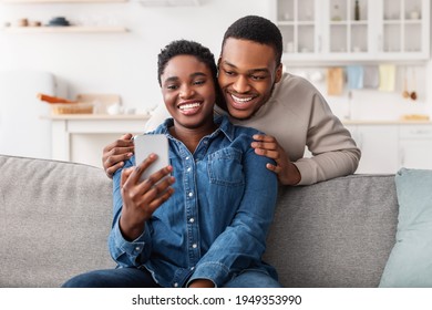 Spending Time Together. Cheerful Black Guy Hugging His Woman From Behind And Looking At Her Mobile Phone. Smiling Lady Sitting On The Couch, Showing Device To Her Man, Watching Photos, Ordering Online