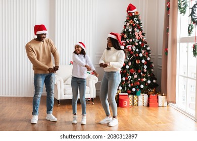 Spending Time Together On Holiday Concept. Cheerful black girl moving and dancing to music with her excited parents in santa hat having fun with mom and dad, enjoying Xmas with family in living room - Powered by Shutterstock