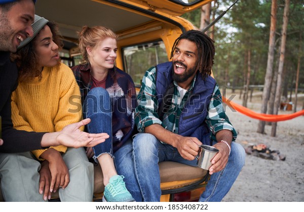 Spending\
time together. Portrait of multiracial young people sitting at the\
car trunk drinking tea and telling funny\
stories