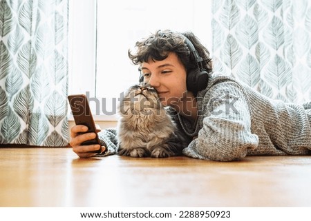 Spending your free time at home with your cat. teenage girl in headphones, with mobile phone, lies on floor in living room near large window, with fluffy Maine Coon cat, listens music