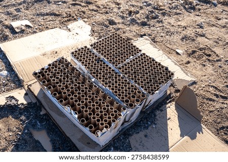 Spent fireworks cake boxes with empty cardboard launch tubes sitting on flattened cardboard on sandy ground, aftermath of pyrotechnic celebration with ash and debris in bright sunlight
