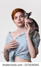 Sphynx Kitten Sitting On Shoulder Of Beautiful Redhead Young Woman 25 Years Old. Portrait Of Female With Short Hair Dressed In Striped White-blue Shirt. Studio Shot On White Background. Part Of Series