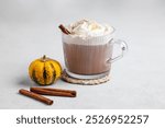 Spice latte or hot chocolate with whipped cream in glass cup on light background with decorative pumpkins and cinnamon stick. Concept of trendy autumn warming drink. Front view and copy space.