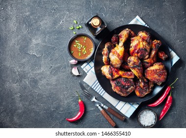 Spicy Grilled Caribbean Jerk Chicken drumsticks and thighs on a black platter on a concrete table with sauce in a bowl, horizontal view from above, flat lay, empty space