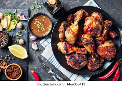 Spicy Grilled Caribbean Jerk Chicken drumsticks and thighs on a black platter on a wooden table with ingredients at the background, horizontal view from above, flat lay, empty space, close-up
