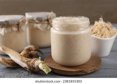 Spicy horseradish sauce in jar and roots on grey wooden table, closeup