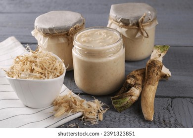 Spicy horseradish sauce in jars and roots on grey wooden table