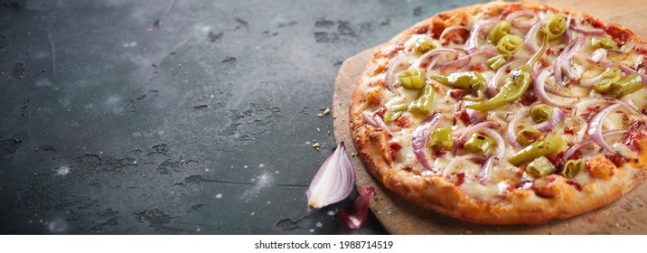 Spicy Italian pizza with jalapeno peppers and red onion trimmings on mozzarella cheese served whole on a wooden cutting board on slate in a panorama banner with copyspace