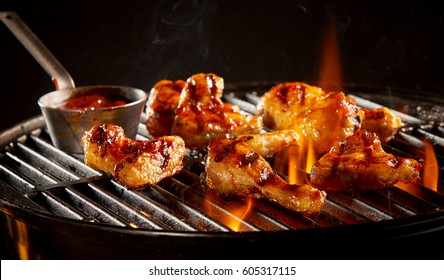 Spicy marinated chicken wings and legs grilling on a summer barbecue with hot flames and a small pot of marinade in a close up view