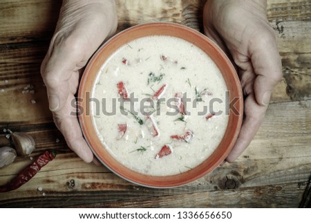 Spicy soup with sour cream, bread and spices on burned wooden table. Traditional rural dinner. Old woman hands holds clay bowl. Toned.