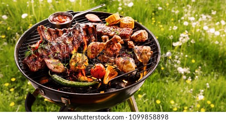 Spicy spare ribs, assorted veggies and chicken drumsticks grilling on a portable barbecue outdoors in a spring meadow with dandelions in a panorama format