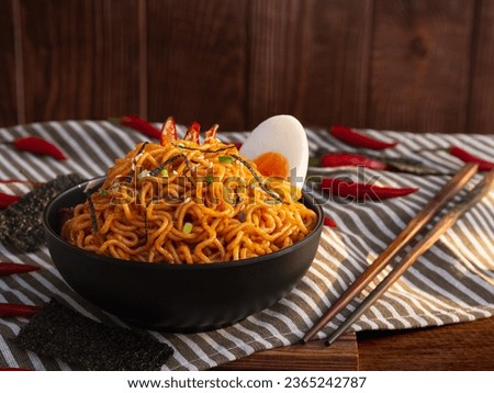 Spicy stir fried instant noodle with boiled eggs, white sesame, seaweed sheets, spring onion in black bowl on wooden table background. Asia food