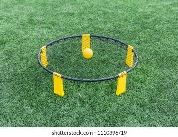 A Spike Ball Game Is Set Up On A Green Turf Field With A Yellow Ball Resting On The Netting. 