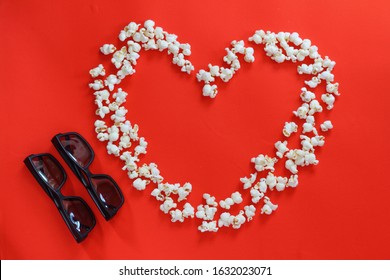 Spilled popcorn in a striped cardboard box and couple 3D glasses on a red background. Popcorn lined as a heart sign. Cinema, movies and entertainment concept. Top view, mock up, copy space.