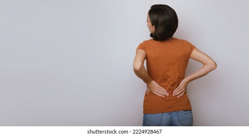 Spinal cord problems on woman's back. Chronic back pain. Young brunette woman is holding her lower back, while standing and suffering from unbearable and severe pain. Spine osteoporosis. - Shutterstock ID 2224928467
