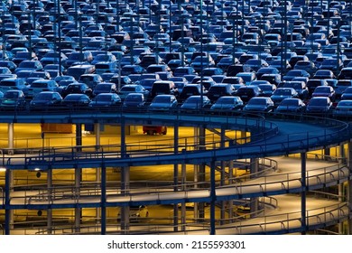 Spiral ascent ramp driveway of big car park in Cologne Germany with yellow illumination. Garage building with steel and concrete construction at blue hour twilight and hundreds of cars on top deck.