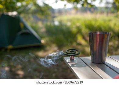 Spiral From Mosquitoes Is Smoking While Standing On A Table Next To A Metal Glass Against The Background Of A Camping Tent Outdoor