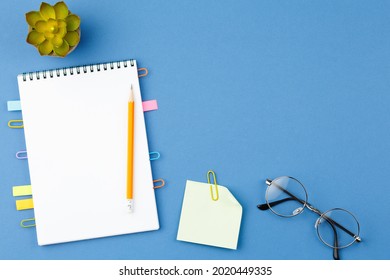 Spiral notebook with bookmarks from paper clips, note sheets, pencil, glasses and potted plant on blue isolated background. Office concept. Top view.
